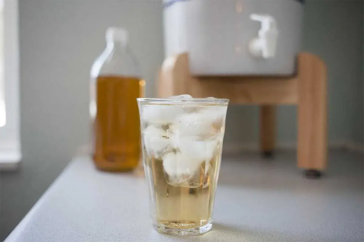 A hand pouring raw unfiltered apple cider vinegar from a bottle into a measuring spoon beside a clean plastic chicken waterer on an outdoor potting bench
